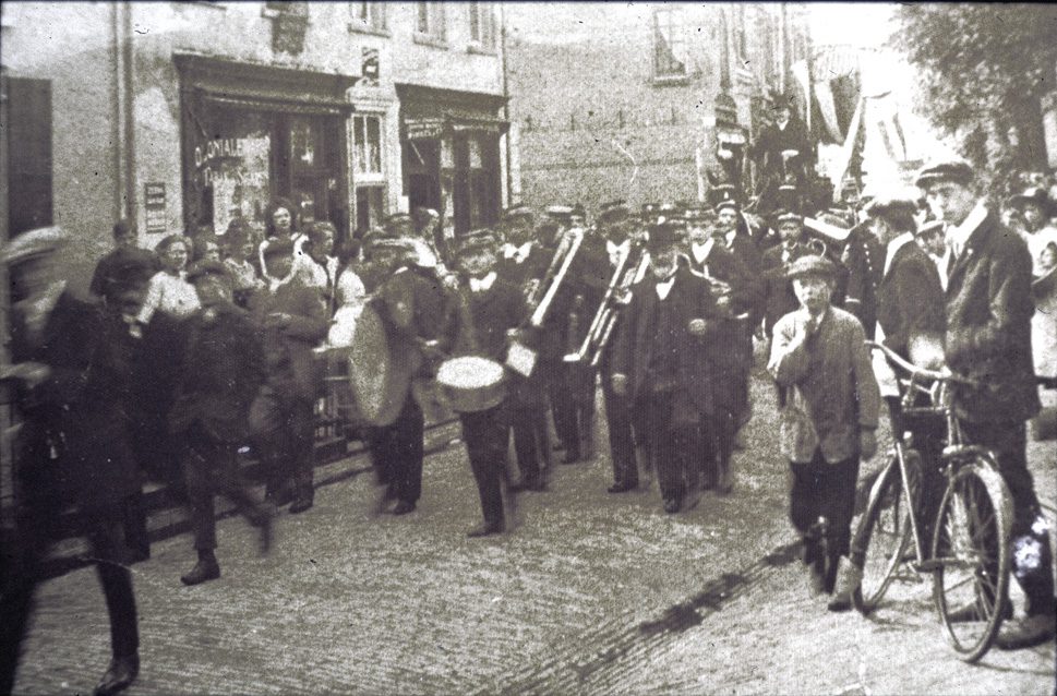 Evert de Graaf, rechts met hoed en baard, temidden van "De Eenvoud". Ze passeren "de Handwerkwinkel", van mej.H. Vlasveld handelend in tricotages, garen- en band, en modeartikelen.